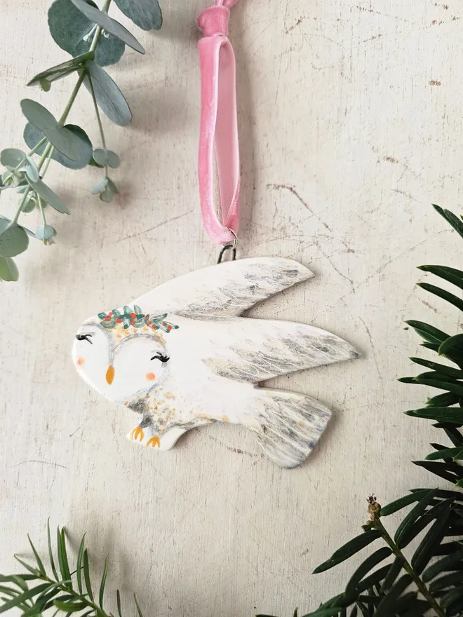 Decorative owl ornament with floral crown hanging from a branch on a soft creme background.