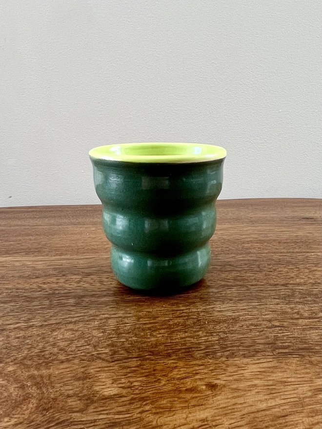 ceramic wavy tumbler with dark green glaze on the outside and lime green on the inside. the tumbler holds 250ml of liquid and sits on a wooden table