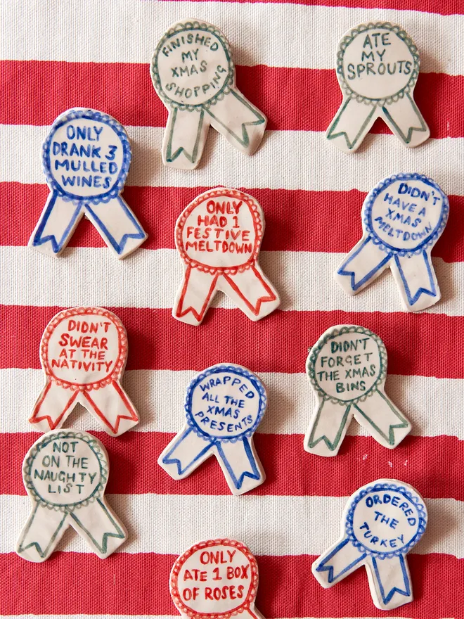 A selection of Red, Green & Blue Handmade Ceramic Christmas Award Badges, handpainted with different phrases, atop a striped red and white tablecloth