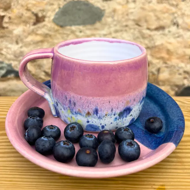 Shoreline Large Round Mug, a colourful mug on a wooden surface against a brick backdrop. It is accompanied by fruits. 