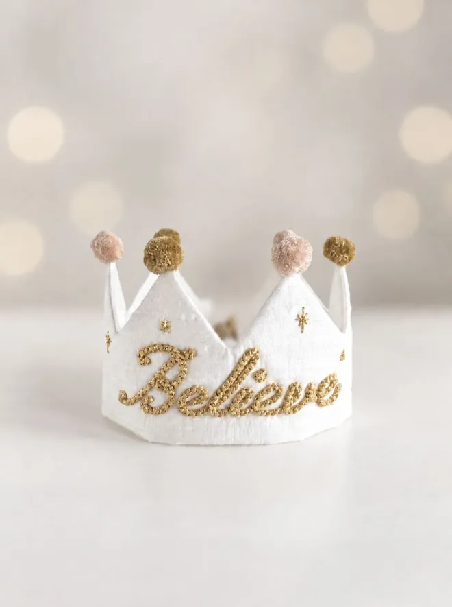Handmade white silk Believe Christmas crown with metallic gold embroidery, gold star details and soft blush and antique gold pom-poms.