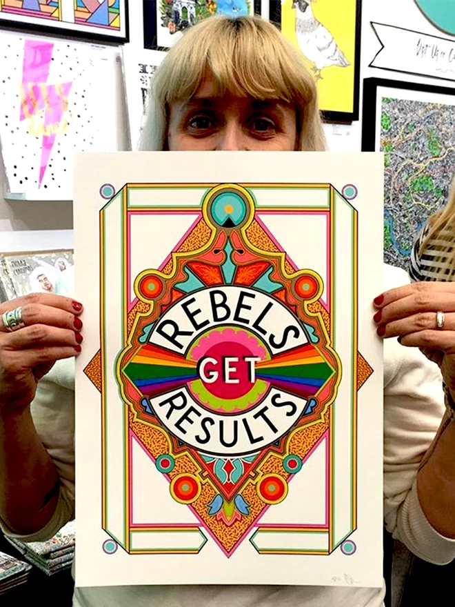 A caucasian woman with a blonde fringe and blue eyes holds the print in front of their face. Rebels Get Results is written in white on a black background at the centre of this vibrant, abstract portrait illustration, with a white background and rainbows emitting from the centre and multi-coloured detailing. 