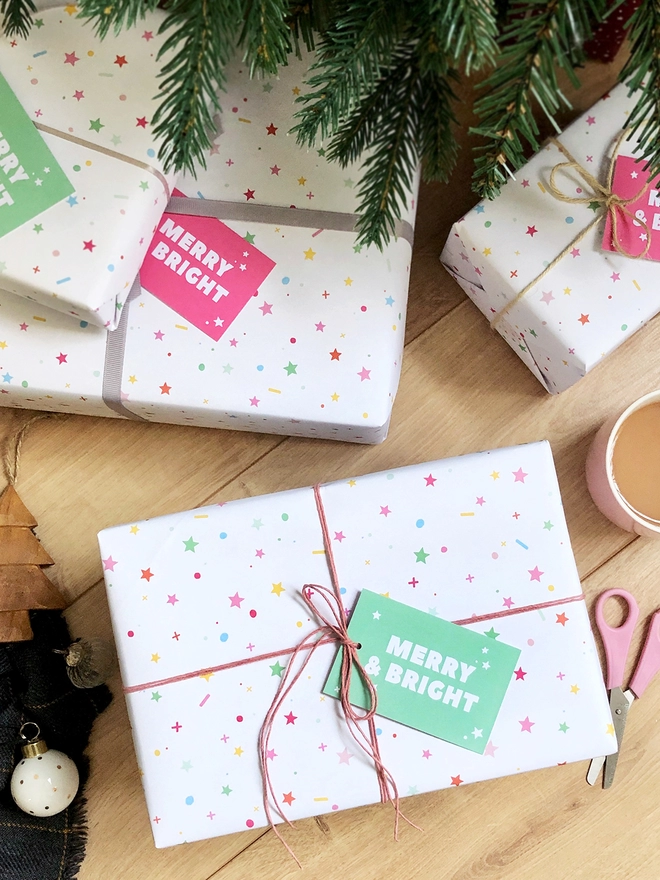 Gifts wrapped in pastel star Christmas wrapping paper are on a wooden floor beneath a Christmas Tree.