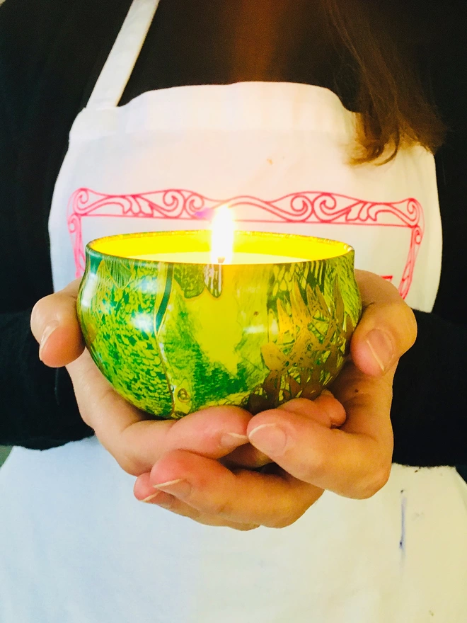 artist holding lit abundance of love gift of light travel charity candle in a tin wearing apron