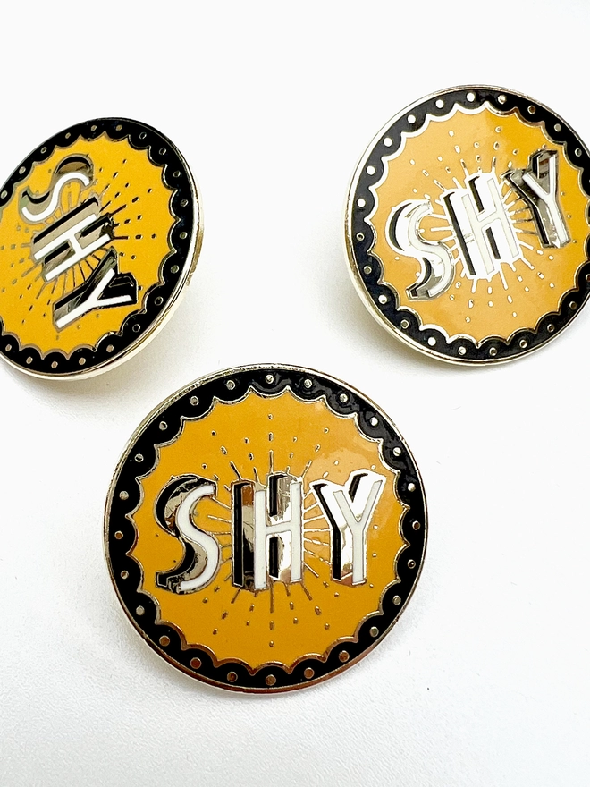 A close up of three SHY cloisonne brooches, they are mustard, black, white and brass coloured. 