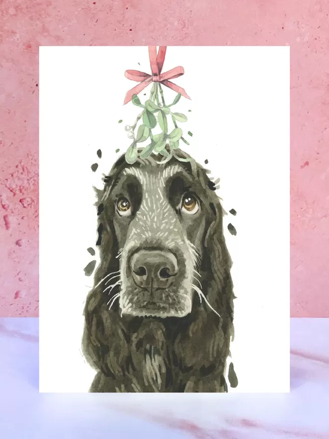 A Christmas card featuring a hand painted design of a Black Cocker Spaniel, stood upright on a marble surface.