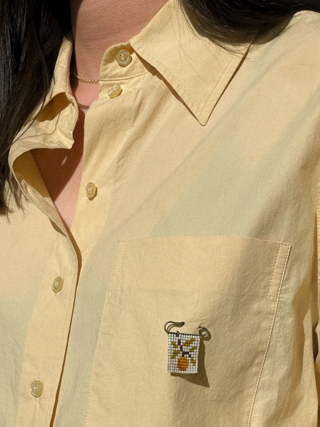 'flora viii' tiled beaded orange pin. a beaded pin badge with an orange tree branch on a pearl background, fasted to a shirt breast. 