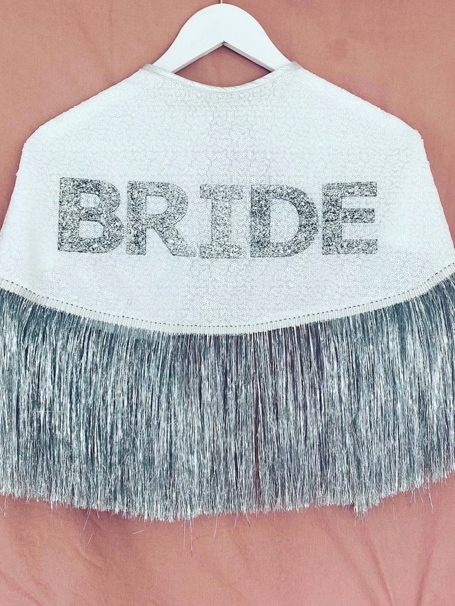 The gretna bride cape on a hanger against a pink backdrop. The cape features the word 'BRIDE' in silver chunky glitter on top of white sequins. The cape has a silver tinsel trim along the bottom,