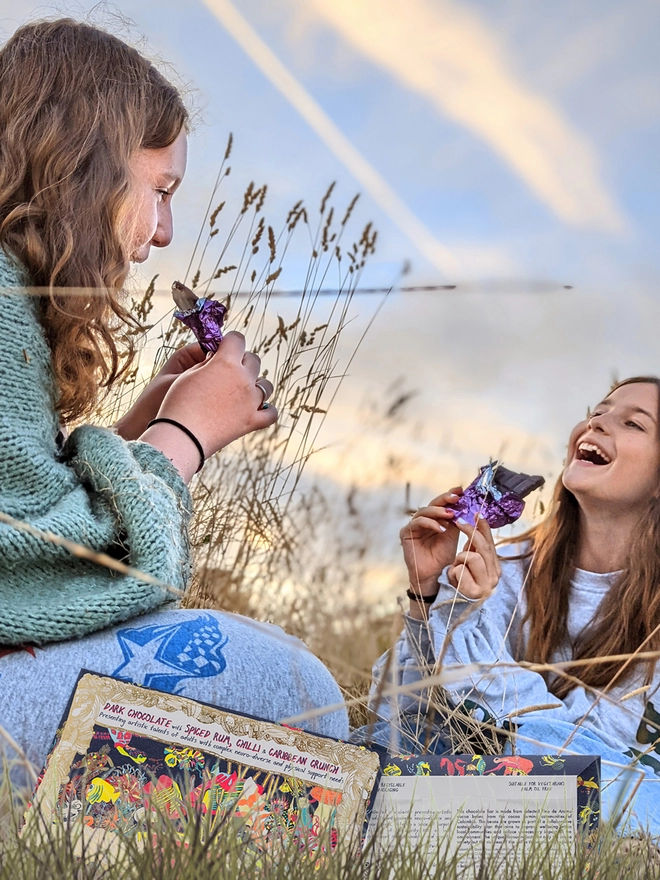 Two happy girls in a field enjoying charity dark chocolate bar wrapped in purple foil 