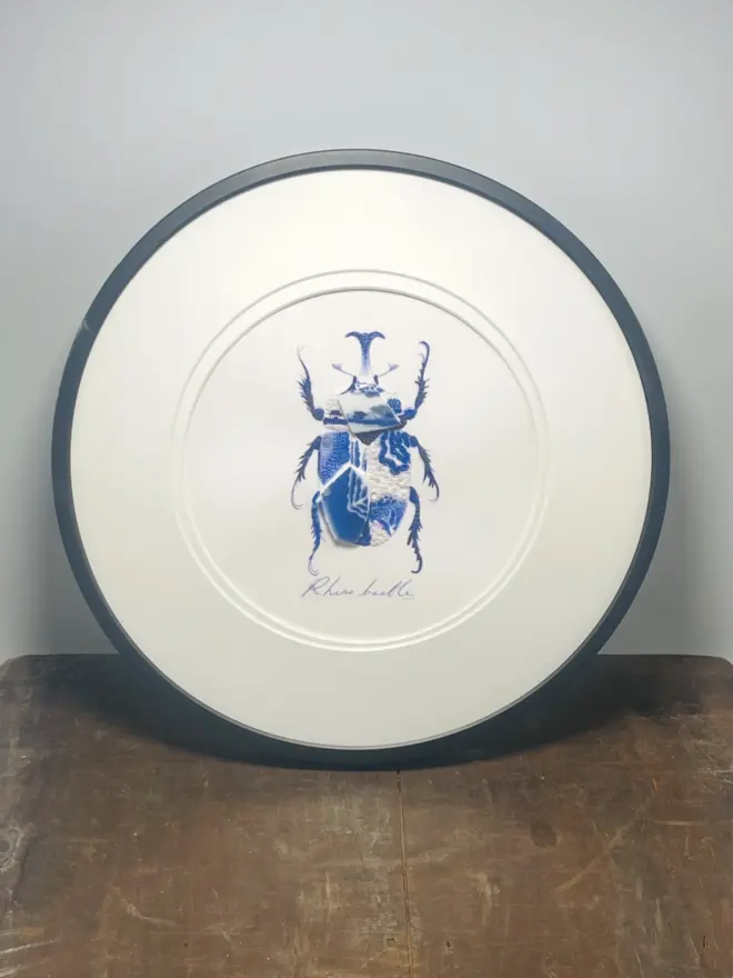 Hand-drawn, painted, mosaic clay, vintage blue Chinese Willow Tree pottery insect on white double mounted card, in a black circular frame.