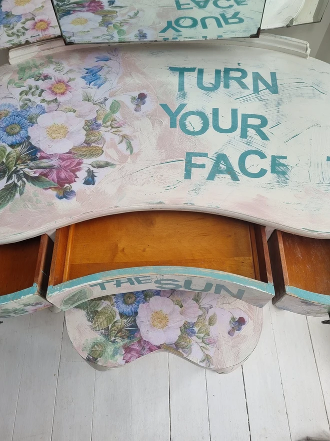 A painted dressing table in white and pale pink with large florals in blues and mauves and text in turquoise that reads 'turn your face to the sun'. The whole piece is distressed in the shabby chic style.