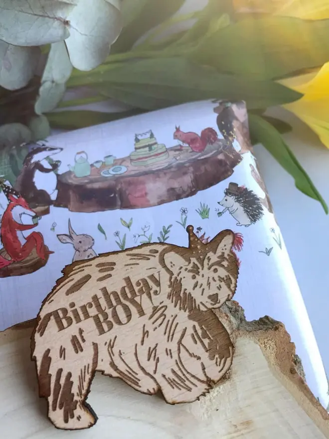 Birthday Boy Wooden Bear Badge, A wooden bear badge engraved with "Birthday BOY!" sitting on a tree slice next to woodland-themed gift wrap and yellow flowers.