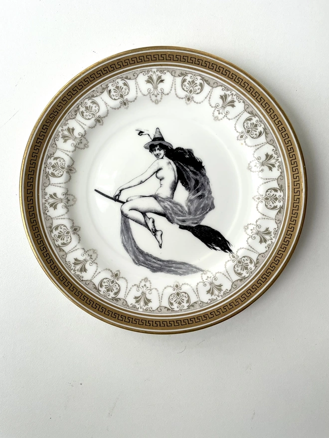 vintage plate with an ornate border, with a printed vintage illustration of a witch on a broomstick in the middle 
