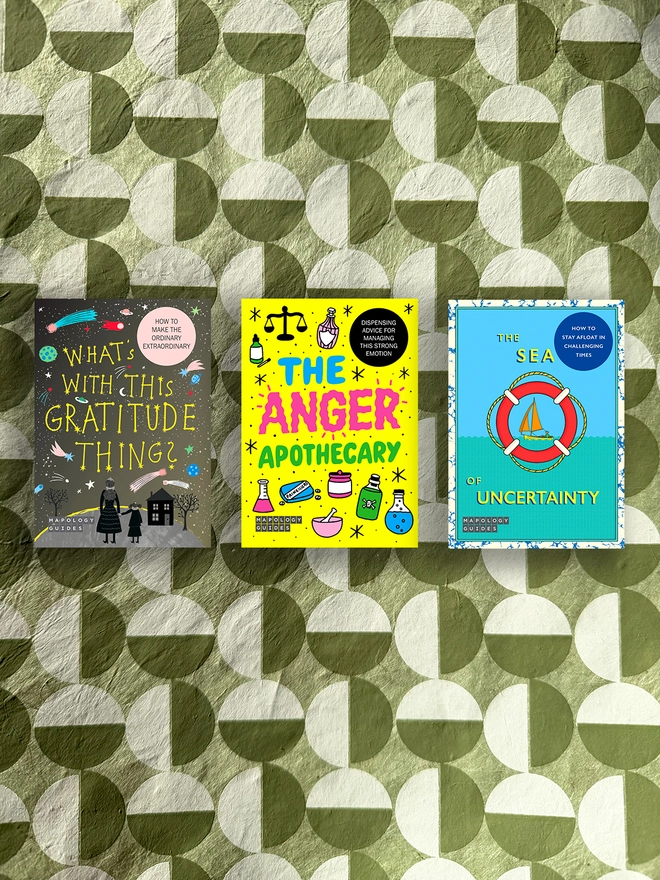 A bundle of three front Mapology Guides covers on a multi-coloured and multi-patterned background. The maps are: “What’s With This Gratitude Thing?” featuring a starry night sky, planets, comets and two figures standing near a house. Text reads “How to make the ordinary extraordinary.”
The Anger Apothecary Mapology Guide cover with bright illustrations of bottles and scales on a yellow background.
The Sea of Uncertainty Mapology Guide cover showing a sailboat inside a red lifebuoy with blue waves and yellow lettering 