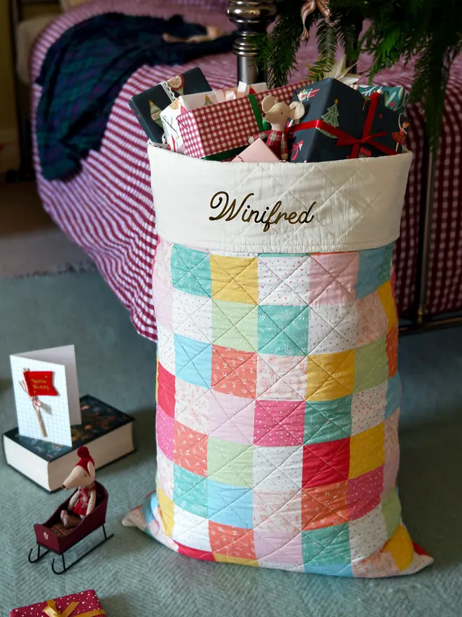 A handmade pastel patchwork Christmas sack stands beside a bed.