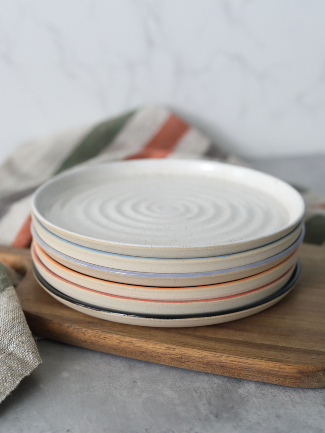 A stack of five handmade ceramic side plates on a wooden board, topped with a speckled white plate showing a concentric swirl texture, with coloured rim edges visible beneath in lavender, mandarin orange, cherry red, and black, styled with a striped linen cloth against a marble background