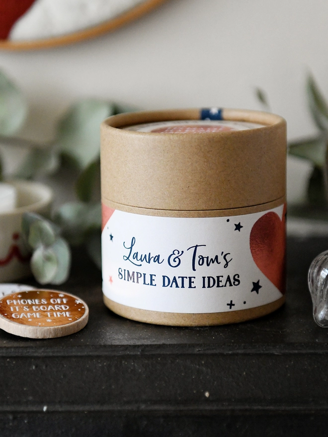 A cardboard jar with a personalised label that read Simple Date Ideas stands on a black mantlepiece in front of gentle greenery.