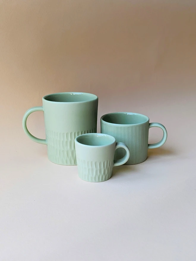 Three handmade porcelain mugs and coffee cups in a Pale Rockpool Green. A large tea mug with a hand carved scalloped texture around the base, a small mug for flat white coffee which is hand carved in a combed texture, and espresso cup with scalloped hand carving around the base. All are glazed inside and matte outside.