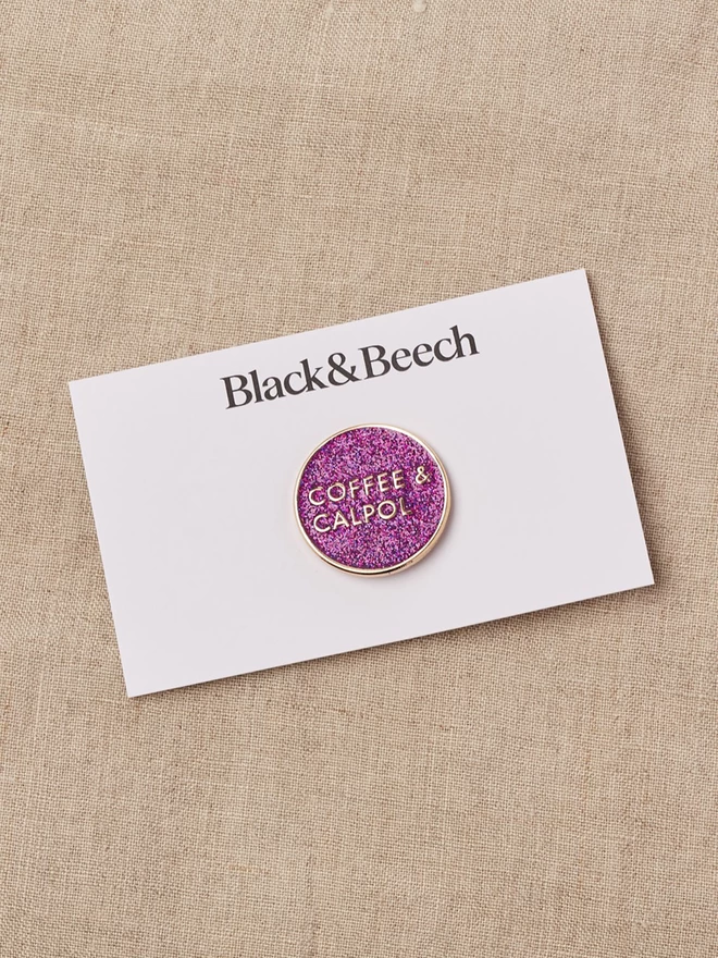 A round purple glitter enamel pin with the words Coffee and Calpol written in gold, on black backing card 