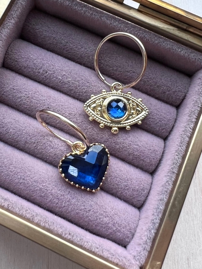 Mismatching gold hoop earrings with faceted blue heart charm and blue and gold evil eye charm inside velvet jewellery box
