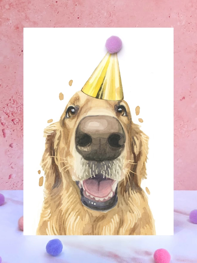 A greeting card featuring a hand painted design of a golden retriever, stood upright on a marble surface surrounded by pompoms. 