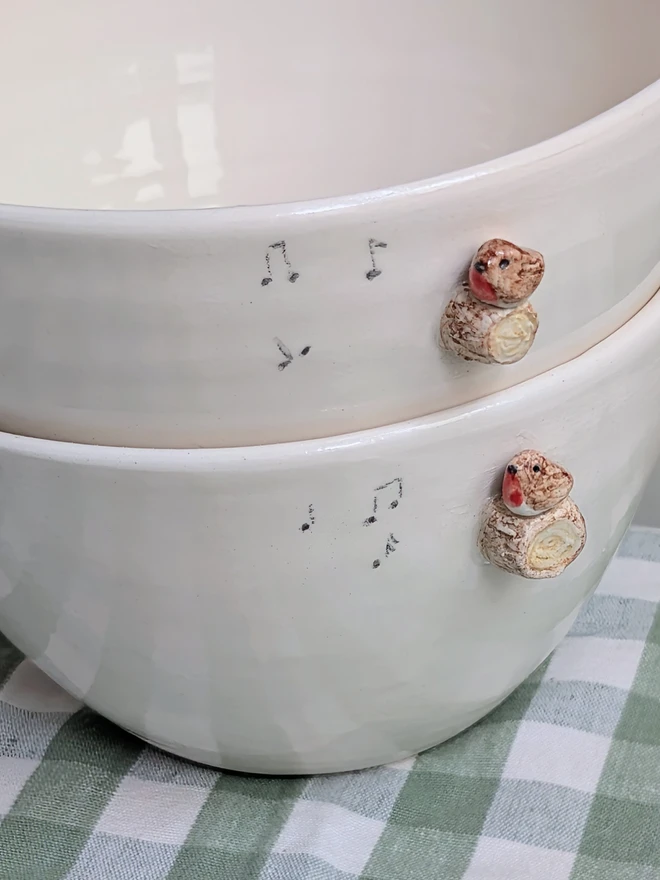 stack of two white ceramic bowls each with a tiny modelled robin perching on a modelled branch with musical notes resting on a green gingham tablecloth 