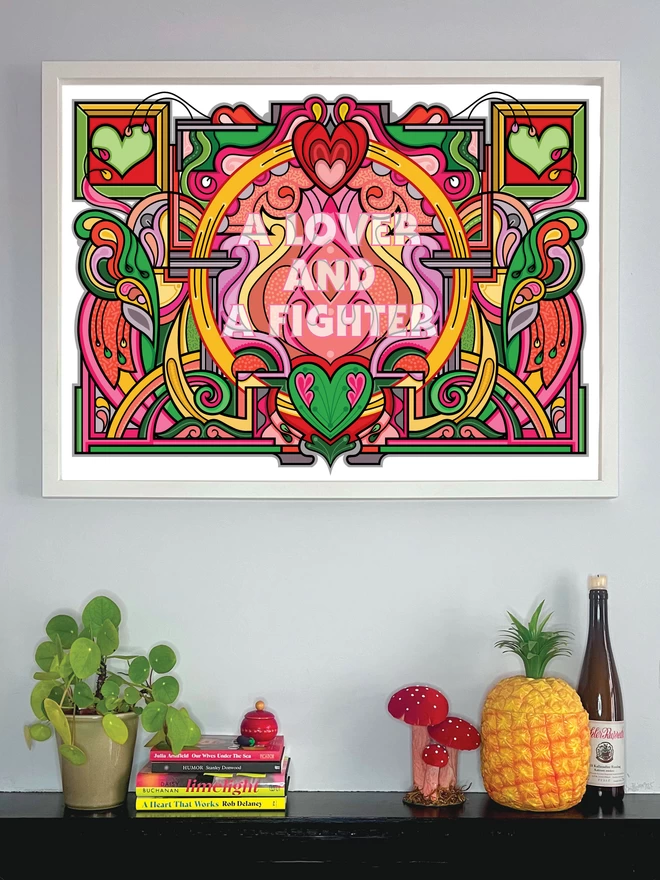 The illustration featuring "A lover and a fighter" faintly written over the colourful, abstract design sits in a white frame above a shelf decorated with a pot plant, books, a toadstool ornament, a plastic pineapple and a wine bottle. 