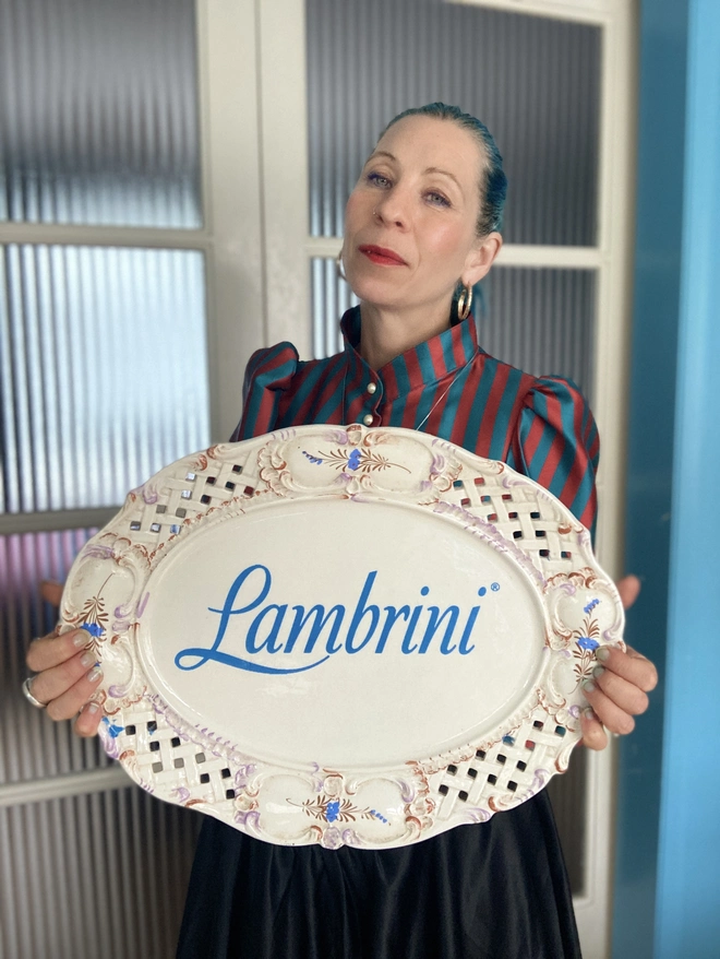 Haus of Lucy holding a large, cream coloured vintage plate with the 'Lambrini'  logo across it in blue.