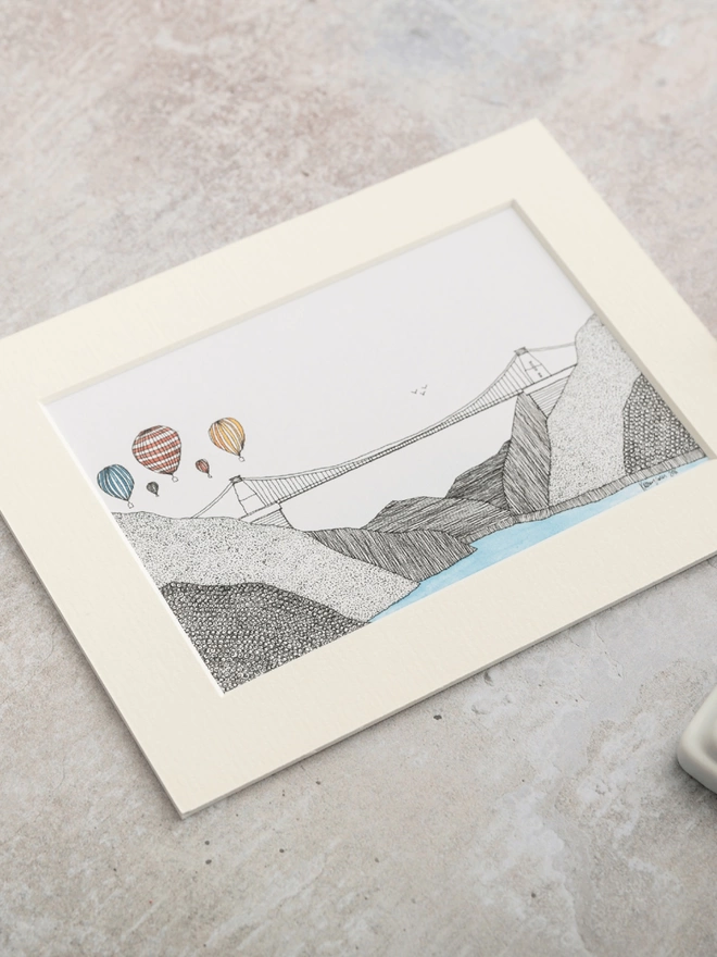 Print of detailed pen and watercolour drawing of air balloons above Clifton Suspension Bridge, in a soft white mount
