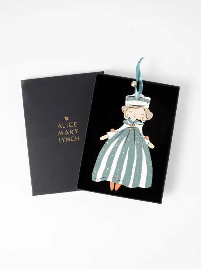 Sailor Queen Tree Dancer. the sailor queen tree dancer hanging ornament sits inside its black presentation box, with the lid open to reveal "alice mary lynch" embossed in gold lettering. the figure has curled hair, gold hoop earrings and a striped sailor hat, wearing a teal and cream striped gown dotted with gold. the branded box gives the piece a polished, gift-ready presentation.