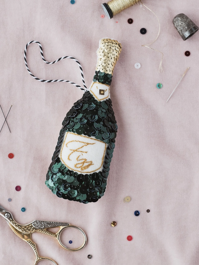 A green sequinned champagne bottle ornament on a pink cloth