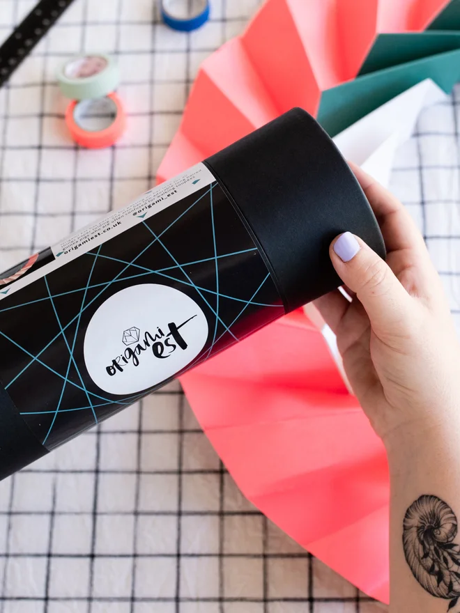 Woman holding an origami kit in a black tube with paper ornament and craft materials in background.