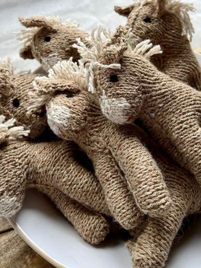 Hand Knitted Natural Dyed Wool And Alpaca Yarn Donkey Gift, a group of hand knitted donkeys sitting in a white bowl on a wooden chopping board on a table cloth. They are accompanied by some hey and some knitting accessories. 