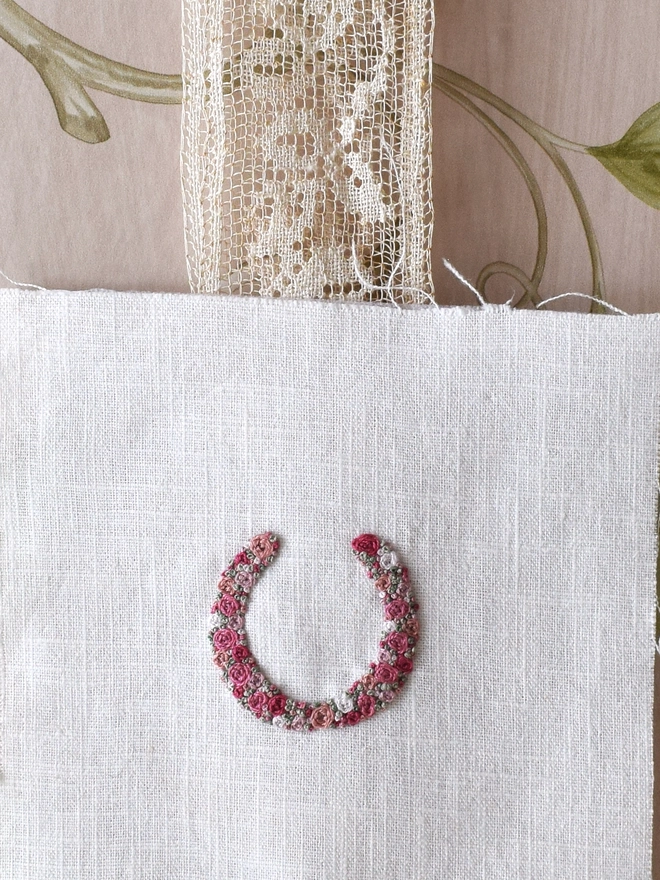 An embroidered Pink Roses Horseshoe, of woven wheel roses in 5 shades of pink with French Knot green grass.  Pinned to a wide lace ribbon.