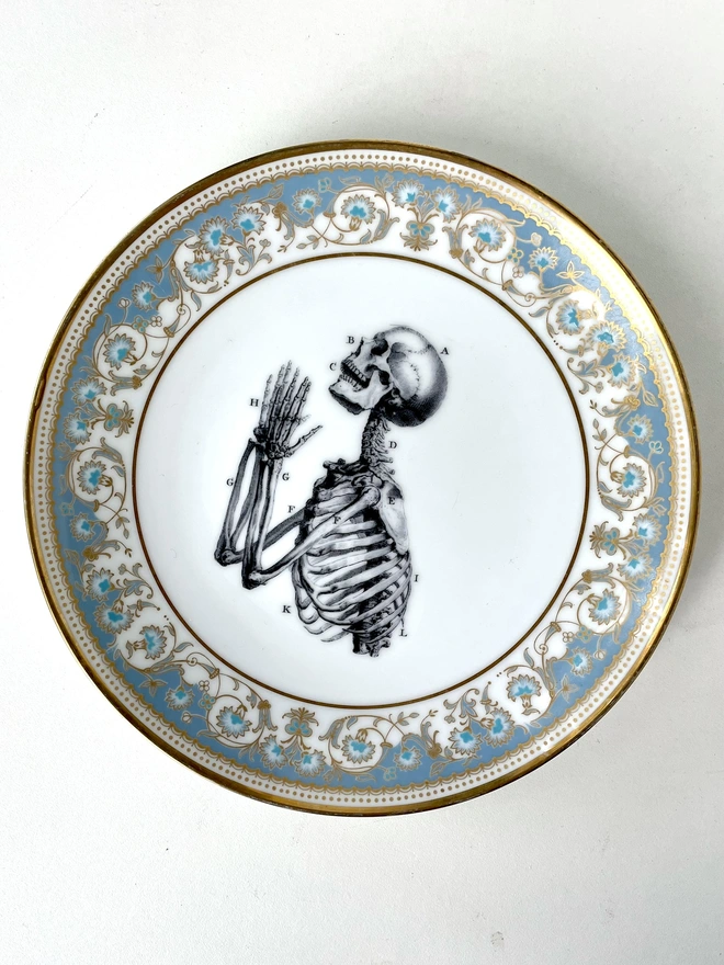 a vintage ornate plate that has a patterned border, and in the centre i have print a black and white illustration of praying hands skeleton in a vintage style