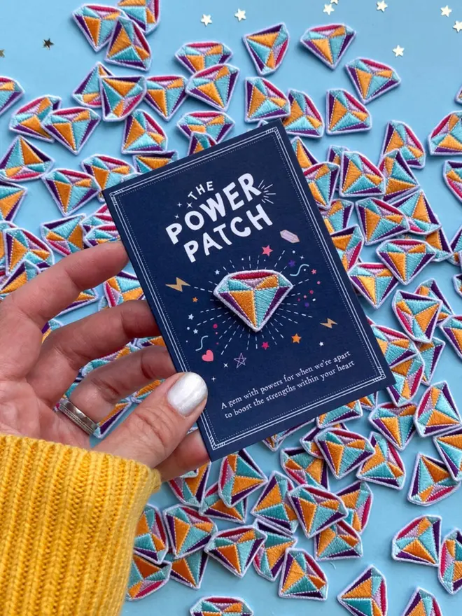A hand holds the navy card with its colourful embroidered gem patch. Piles of matching patches and gold confetti stars fill the background.