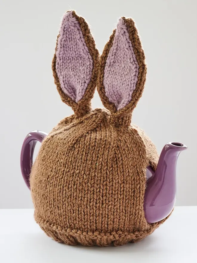 Hand Knitted Bunny Rabbit Tea Cosy, a rabbit bunny tea cosy sitting on a grey surface against a white backdrop. It fits over a purple tea pot. 