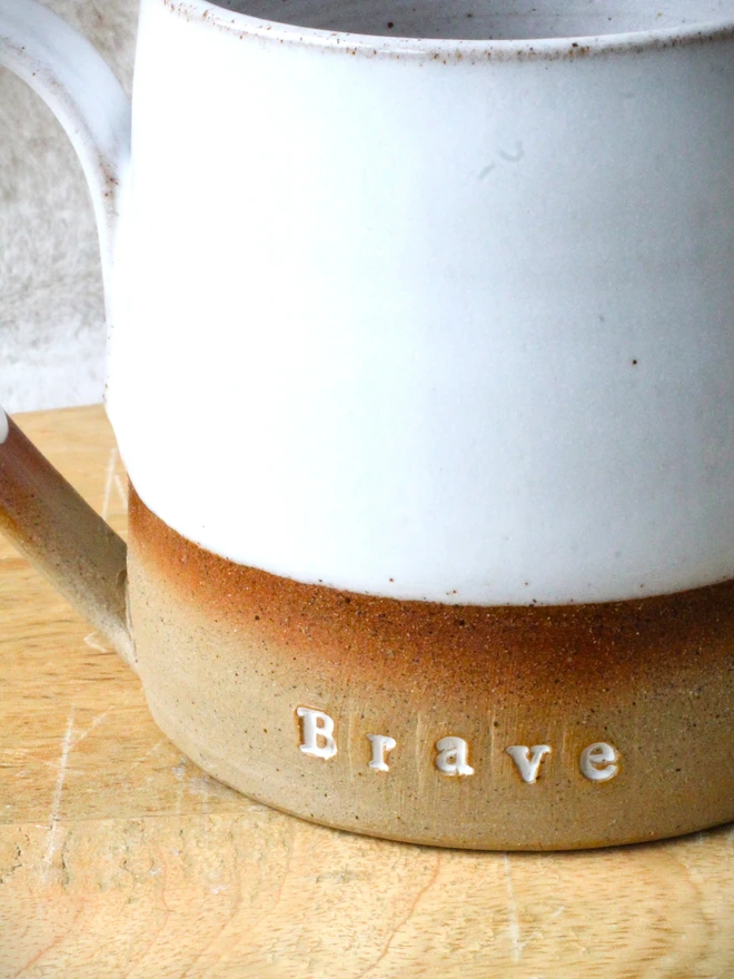 Rustic ceramic mug with a white speckled upper glaze and brown lower half, featuring the word “Brave” on the front, sitting on a wooden table against a neutral background.