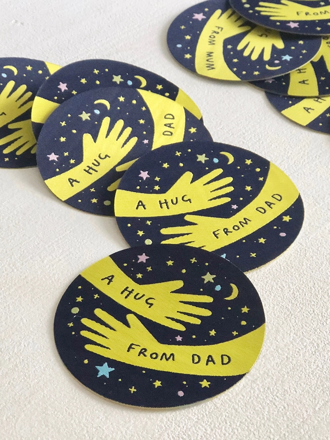Several navy blue woven patches with a yellow hugging arm design and the words "A hug from Dad" are scattered on a white desk.