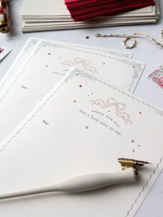 A close-up of cream lined Christmas notelets with a pink ribbon bow, scattered red and silver stars, silver scrollwork corners and the words "written with love, just a little note to say…". A white calligraphy pen rests on top, with red and cream envelopes and twine behind.
