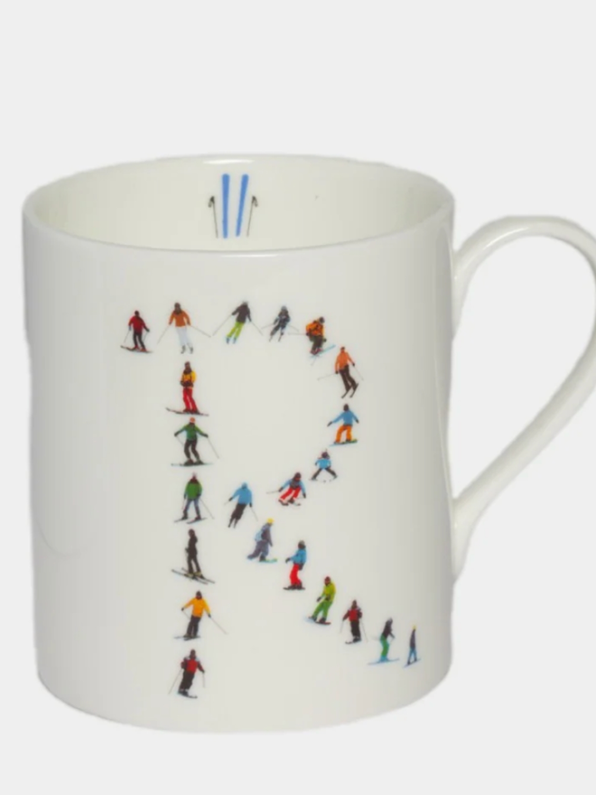 A simple white mug illustrated with an ‘R’ made of small, multicoloured skiers. The mug is set against a plain, white background.