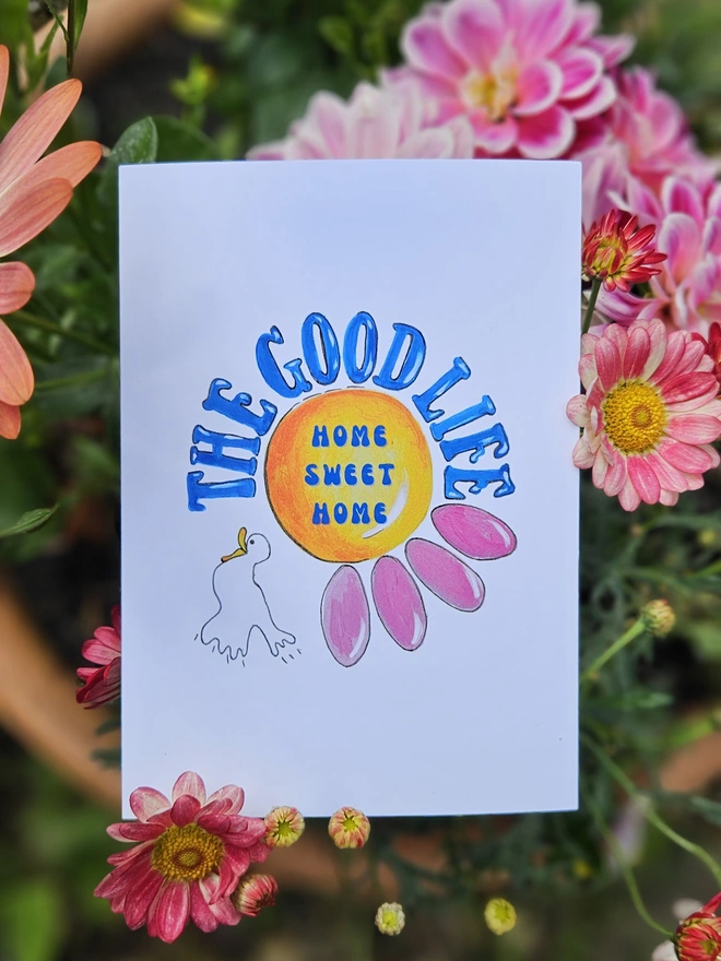 A white card with an illustration of a pink flower and a white bird with The Good Life is written around the edge of the flower using a retro font in blue. In the centre of the flower is says in a blue retro font Home Sweet Home 