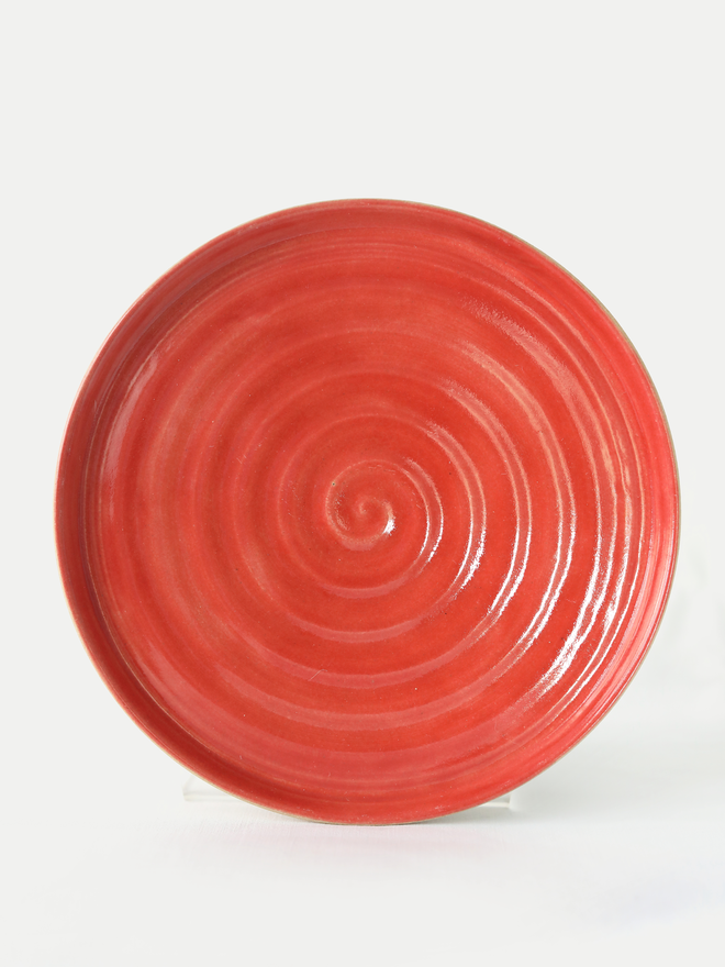 A handmade ceramic side plate in cherry red glaze, photographed from the front against a plain white background, showing a natural concentric swirl texture from the wheel-throwing process
