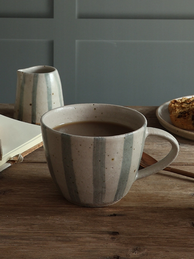 Handmade Ceramic Stoneware Mug in Blue and White Stripe sat on a wooden surface alongside a small handmade jug in the same style and cookies sat on a handmade ceramic plate.   