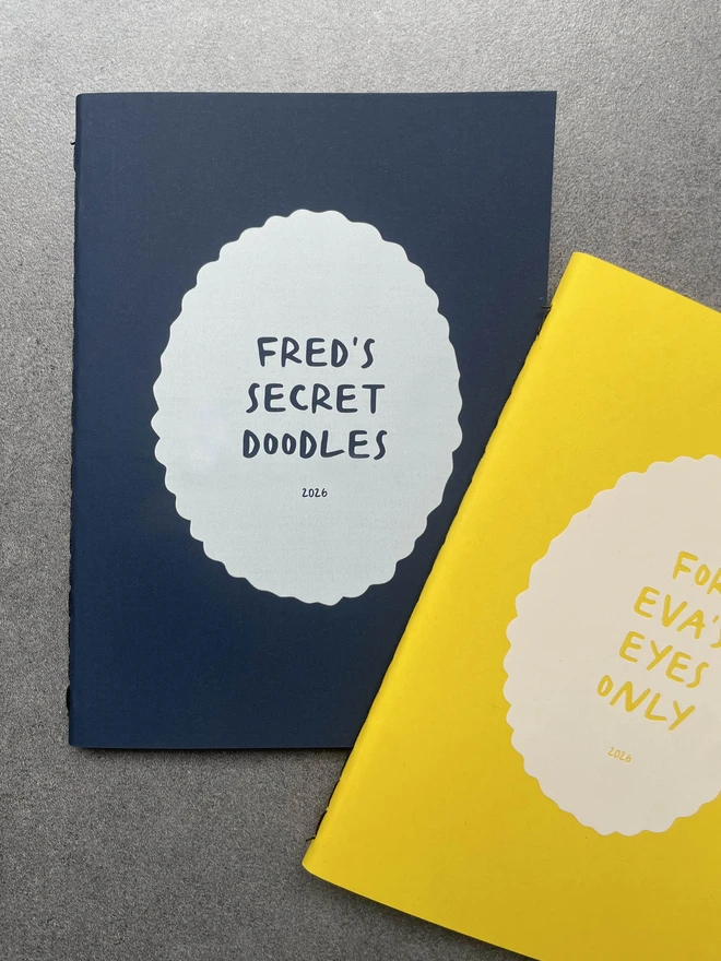 two personalised notebooks. One is a dark navy and says "Fred's secret doodles" and the other biright yellow book says "For Eva's Eyes only" 