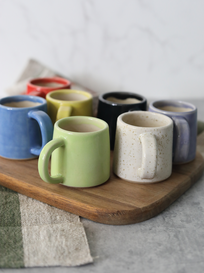 Eight handmade ceramic espresso mugs grouped on a wooden board, showing the full colour range of spring green, speckled white, blue, yellow, black, purple, and red, each with a chunky loop handle and containing espresso, styled with linen cloths against a marble background