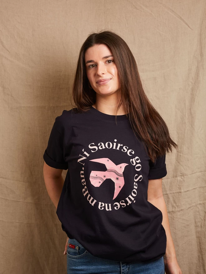 Model is shown wearing a Black & Beech navy heavyweight t-shirt with the words Ní Saoirse Go Saoirse Na mBan in pink written in a circle surrounding a pink bird