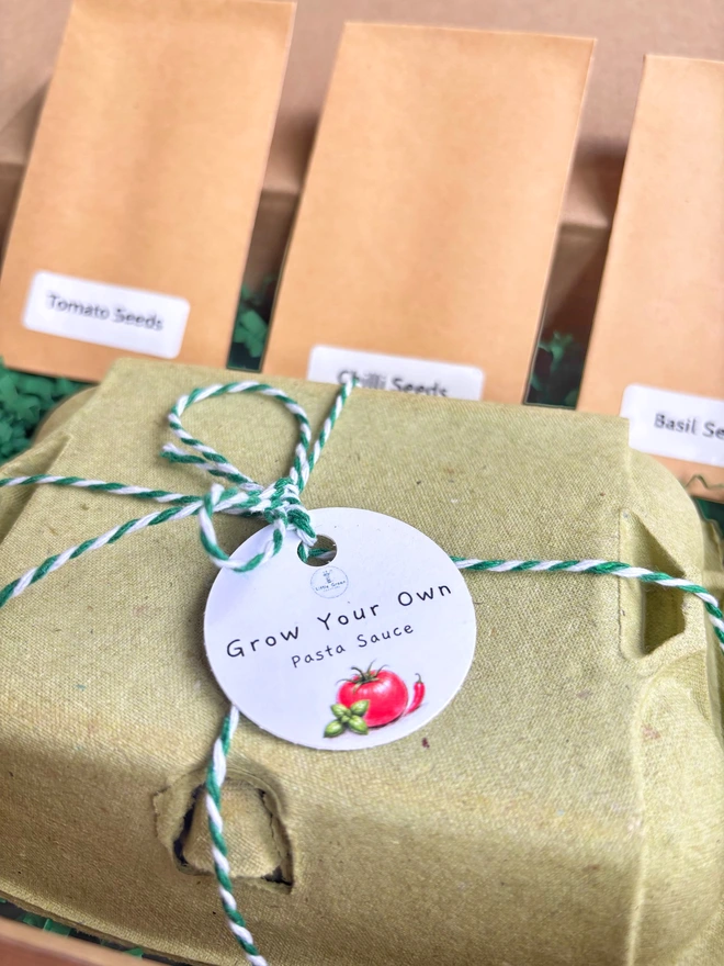 closed green egg box tied with green string and a grow your own tomatoes tag. Behind are three kraft seed packets and dark green paper box filller