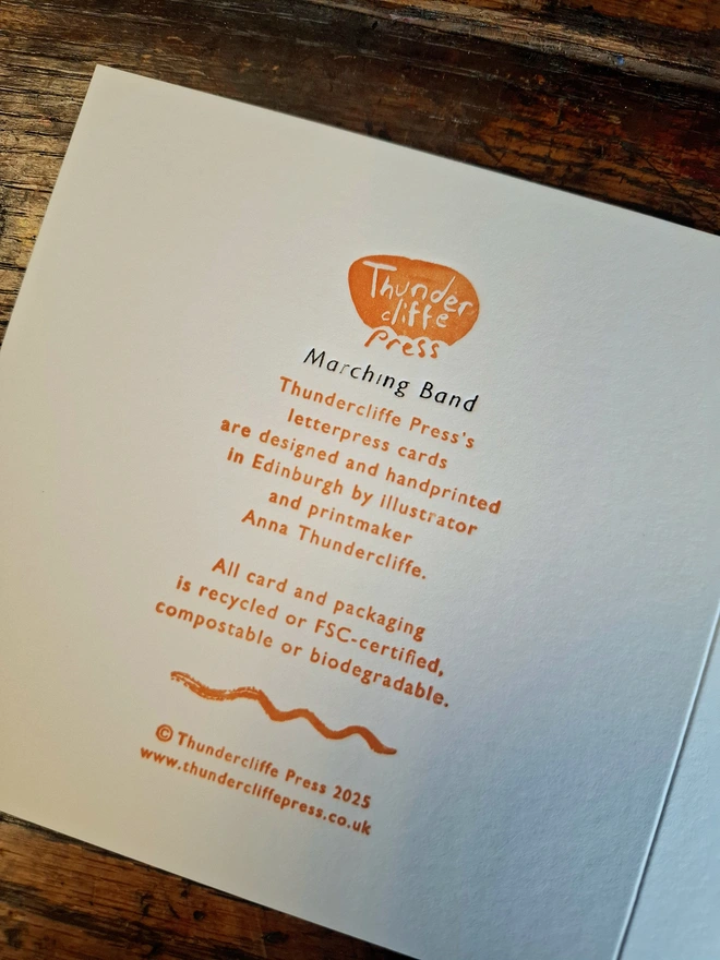 The reverse of a greetings card, showing a logo and text, lying flat on a wooden surface.
