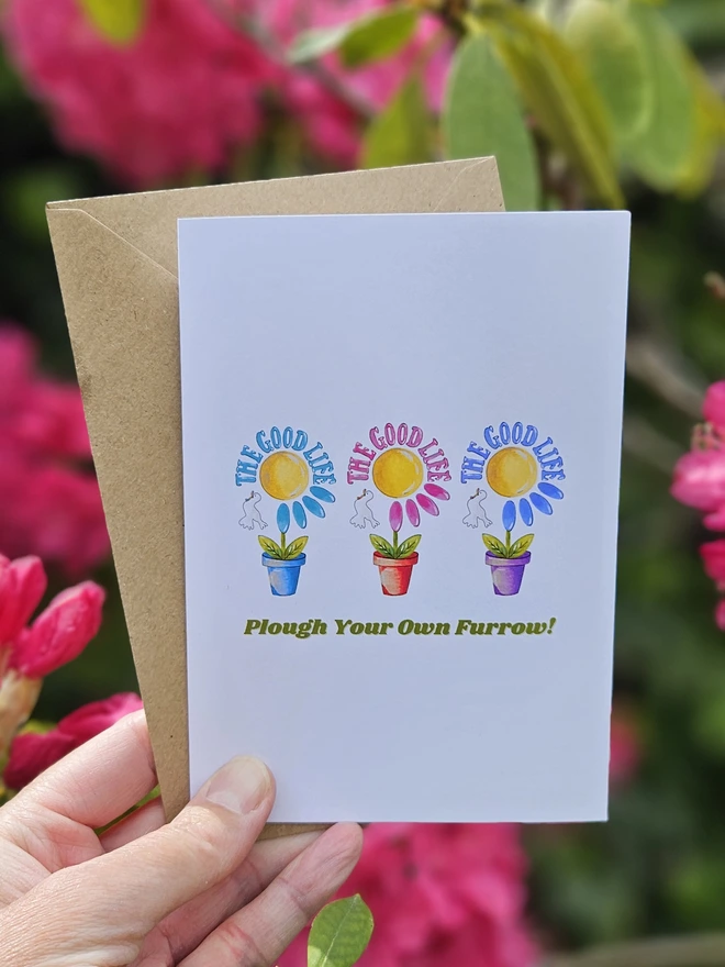 A white card with an illustration of  3 colourful flowers each in a terracotta pot displayed in a row with a white bird. The Good Life is written using a retro style font in 3 colours. It says at the bottom of the card Plough Your Own Furrow!